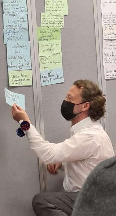 Torben Klußmann hockt vor einer grauen Pinnwand und trägt eine FPP2-Maske während er bunte Moderationskarten an die Wand anpinnt.