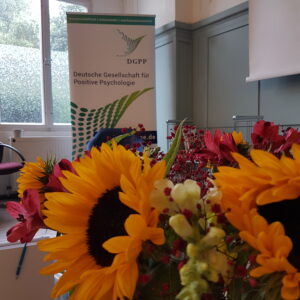 Sonnenblumen in einem bunten Strauß gebunden im Bildvordergrund vor einem Aufsteller der DGPP - Deutschen Gesellschaft für Positive Psychologie in deren Seminarraum.