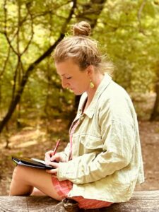 Sabina Grbo sitzt auf einem Baumstumpf seitlich und schreibend im Grünen